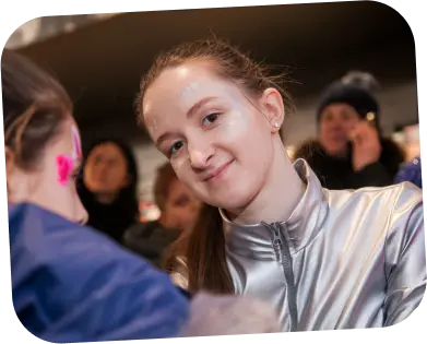 The photograph captures a smiling young woman with a clear complexion and her hair pulled back neatly, engaged in face painting. She is wearing a silver metallic top, giving off a modern and perhaps festive vibe. Her friendly and confident expression suggests she enjoys the activity and interaction. The person receiving the face paint is turned away from the camera, showing only a glimpse of pink design on the side of their face, which looks to be a vibrant, playful pattern. The background is filled with people, indicating the photo was taken at a social gathering or public event where face painting is part of the entertainment. The overall atmosphere seems lively, with the focus on the face painter’s pleasant demeanor as she performs her artistry.