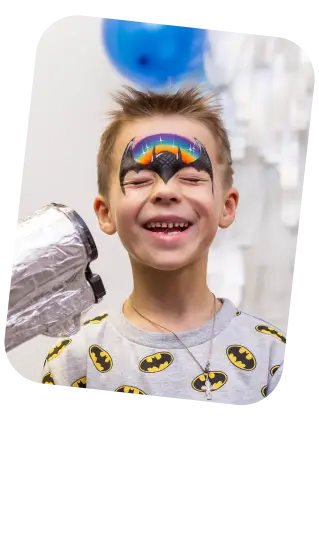 In this joyful image, a young boy is seen with his eyes closed and a wide smile, giving off an expression of delight. He has a colorful face painting over his eyes, featuring a rainbow with clouds on the edges and a superhero emblem in the center, resembling a mask. He's wearing a light grey T-shirt with multiple superhero logos printed on it. A silver foil balloon is partially visible in the top left corner, suggesting he may be at a themed party or a celebration. The background is intentionally blurred, focusing the viewer’s attention on the boy's face and his face painting. His cheerful demeanor suggests he is having a good time, possibly celebrating a special occasion.