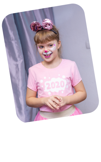 The photo features a young girl with a friendly and excited smile. She is dressed in a playful outfit with a pink T-shirt that has "2020" printed on it, suggesting the photo was taken during or around that year. The T-shirt is paired with a light pink tutu, and she is accessorized with a pink sequined bow headband, reminiscent of a popular animated mouse character. On her face, there is a creative face painting design around her right eye, depicting a whimsical, colorful character with a patch over one eye, adding to the joyful and party-like atmosphere of her look. The girl’s hands are clasped together in front of her, and her stance suggests she is posing for the photo, proud of her festive attire and makeup. The grey curtains in the background imply an indoor setting, possibly a party or celebration where face painting is part of the fun.