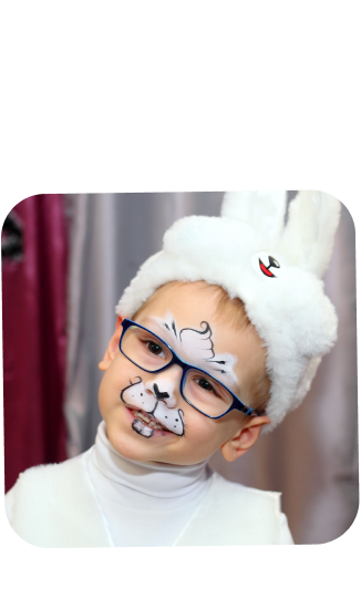 This image captures a young boy with a playful and cheerful expression, wearing a white fluffy bunny hat complete with rabbit ears and a small red detail that mimics the look of a bunny's inner ear. He is also wearing glasses, which adds to his endearing look. The face paint on his nose and cheeks resembles a bunny's snout and whiskers, enhancing the overall theme of his costume. The boy's attire suggests that he is dressed up for a festive occasion, such as a costume party or Easter celebration. His white turtleneck complements the bunny theme, making him look cozy and in character. The backdrop includes a glimpse of a maroon curtain, indicating an indoor setting. His expression and costume create a sense of joy and lighthearted fun.