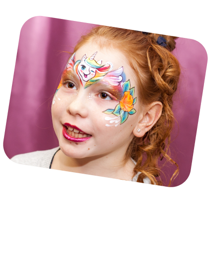 The image depicts a young girl with an expression of joy and slight surprise, possibly in mid-sentence or reaction to something amusing. She has an intricate face painting that sweeps across her right cheek, showcasing a colorful unicorn with a flowing mane, complemented by a vibrant flower and artistic flourishes that give the design a dynamic and whimsical feel. Her hair is styled in a casual updo with loose strands, and she is wearing a white top with visible sparkle, suggesting a playful and festive atmosphere. The background is a soft pink, which harmonizes with the face paint's palette and enhances the cheerful and magical theme of the image. The girl's sparkling eyes and the lively colors of the face paint suggest she is at a celebration, such as a birthday party or a costume event, where face painting is part of the entertainment.
