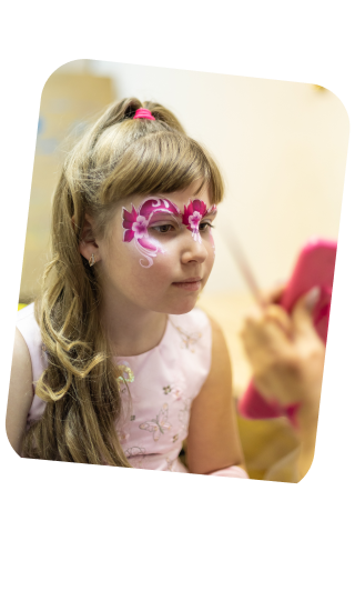 The image depicts a young girl with a contemplative look, gazing at her reflection in a handheld mirror. She has a charming face paint design of pink flowers and swirls around her eye, evoking the image of a floral masquerade mask. Her blonde hair cascades in soft waves over her shoulders, and she wears a delicate pink dress with floral embroidery, enhancing her whimsical, dressed-up appearance. The setting seems to be indoors, with a warm yellowish hue in the background, suggesting a cozy atmosphere, possibly at a birthday party or a dress-up event. The face paint, her outfit, and her absorbed expression all contribute to a sense of childhood wonder and the playful spirit of dressing up and imaginative play.
