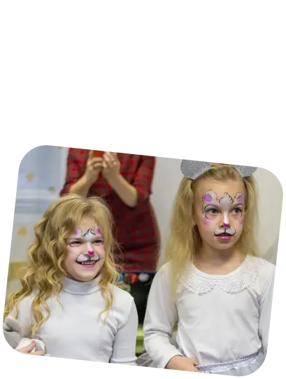 The photo features two young girls with face paint, looking slightly off-camera, with expressions of happiness and a bit of surprise. The girl on the left has a vibrant pink and white face paint design with swirls and dots that suggest a festive, whimsical theme, possibly a fairy or butterfly. She has curly blonde hair and is wearing a light grey or white top. The girl on the right has a similar style of face paint in purple and pink hues, with added elements like a heart on her forehead and detailed designs around her eyes, which may suggest a princess or magical creature theme. She also has blonde hair and is wearing a white top with lace or a delicate pattern at the collar. In the background, an adult with a red top appears to be clapping, adding to the atmosphere of celebration or performance, perhaps at a party or a children’s event.