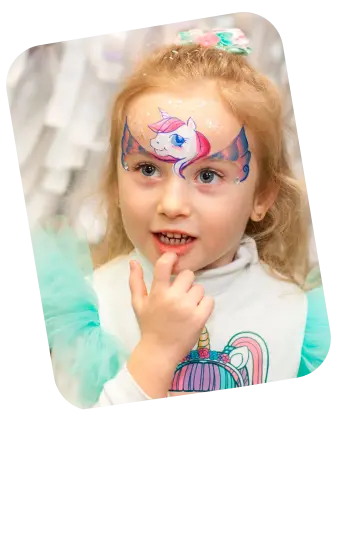 The image shows a young girl with a look of wonder, her hand thoughtfully placed on her chin. She has a colorful face paint design across her forehead, featuring a unicorn with shades of blue and pink, complete with a horn and what appears to be a mane adorned with flowers. Her blonde hair is pulled back from her face, displaying the intricate face paint. She's wearing a white top with a unicorn graphic, suggesting a fondness for this mythical creature. The background is blurred, with white and grey tones that keep the focus on the girl's face. Her expression and the face paint suggest that she's at a festive event, such as a birthday party or a fair, where face painting is part of the entertainment.