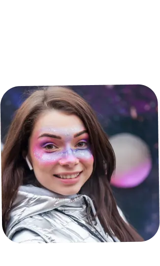 The photograph depicts a young woman with a joyful expression, sporting whimsical makeup that gives off a festive or cosmic vibe. She has sparkling pink and purple hues around her eyes, extending to her temples, with glitter that catches the light, suggesting a starry or galactic theme. Her long brown hair frames her face, and she's wearing a metallic silver jacket, which complements the futuristic look of her makeup. The background is out of focus, with deep purple and black tones and what appears to be bokeh light effects that resemble distant stars or city lights. Her appearance and the background suggest she might be at a themed event, perhaps a music festival or a New Year's celebration.