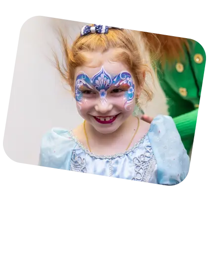 The image features a young girl with a beaming smile, her hair in playful pigtails, accented by a blue hairband with a decorative bow. Her face is adorned with elaborate face paint in shades of blue and white, detailed with swirls and glitter, reminiscent of a butterfly design that covers her eyes like a mask. She is wearing a costume with a blue, possibly princess-themed dress that has silver patterns and sequins, enhancing the magical feel of her attire. The blurred background and her attire suggest she may be at a costume party, a festive celebration, or a performance where children are dressed in fantasy outfits. Her joyful expression and the creative face paint contribute to the lively and enchanted atmosphere of the event.