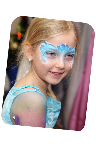 This image shows a young girl with a side profile view, looking off to the distance with a gentle smile. She has a whimsical face painting around her eyes, in shades of light blue and white, designed to resemble perhaps clouds or waves with swirls, which gives her a dreamy, fairytale-like appearance. Her blonde hair is neatly combed, and she's wearing what appears to be a costume with a similar light blue color, suggesting a princess or fairy character theme. The blurred background hints at an indoor setting with warm lighting, possibly a room decorated for a party or festive occasion, as indicated by the glimmers of a Christmas tree or decorations. The overall impression is one of innocence and enchantment, fitting for a child's imaginative play or a themed event.
