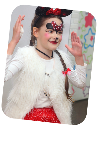 The photo presents a young girl in a playful pose with her hands up as if dancing or performing. She has a creative face painting that resembles a popular animated character, with a red and white polka dot bow, black ears, and a nose to match. Her costume includes a white faux fur vest over a white long-sleeve top and a sparkly red skirt, completing the themed outfit. Her hair is styled in a long braid adorned with a red bow, coordinating with the character-inspired face paint. The setting appears to be an indoor space with artwork in the background, suggesting a school event, birthday party, or children's entertainment venue where she is actively participating in the festivities. Her joyful demeanor captures the essence of childhood fun and imaginative play.