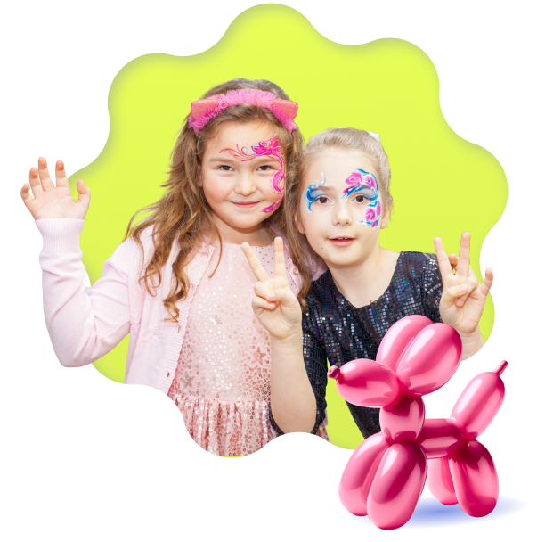 In the image, there are two young girls at what appears to be a festive event or party. Both are smiling and striking poses for the camera; one is waving with an open hand as a greeting, and the other is making a peace sign. Their faces are adorned with intricate, colorful face paint—one in shades of pink and white, the other in blues and pinks—adding to their cheerful and festive appearance. They are dressed in casual yet celebratory attire, one in a glittery pink dress with a matching headband, and the other in a dark long-sleeved top. In the foreground, there is a balloon animal, twisted in the shape of a dog or another quadruped, adding a playful element to the scene. The background is an abstract blend of yellow and green hues, creating a blurred effect around the subjects.